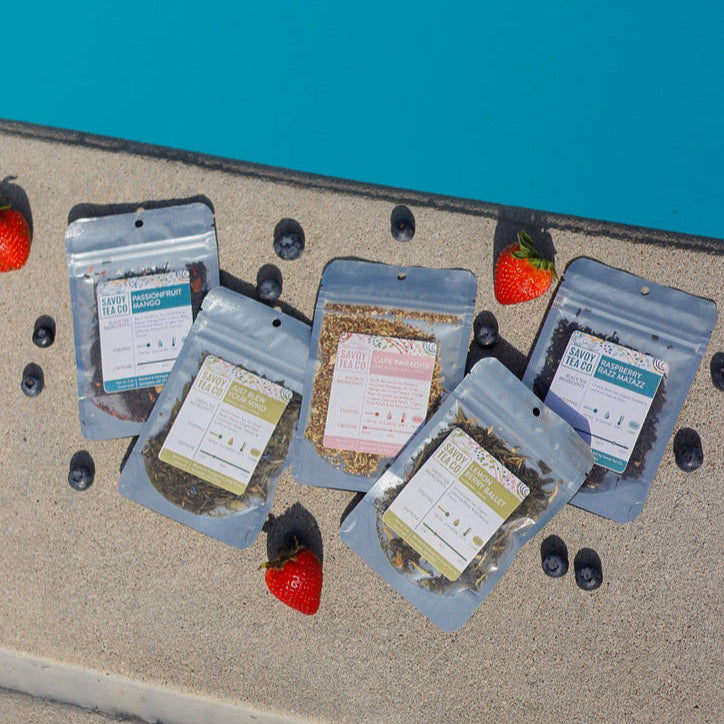Five packages of loose leaf tea on a textured surface next to a pool with strawberries and blueberries around them.