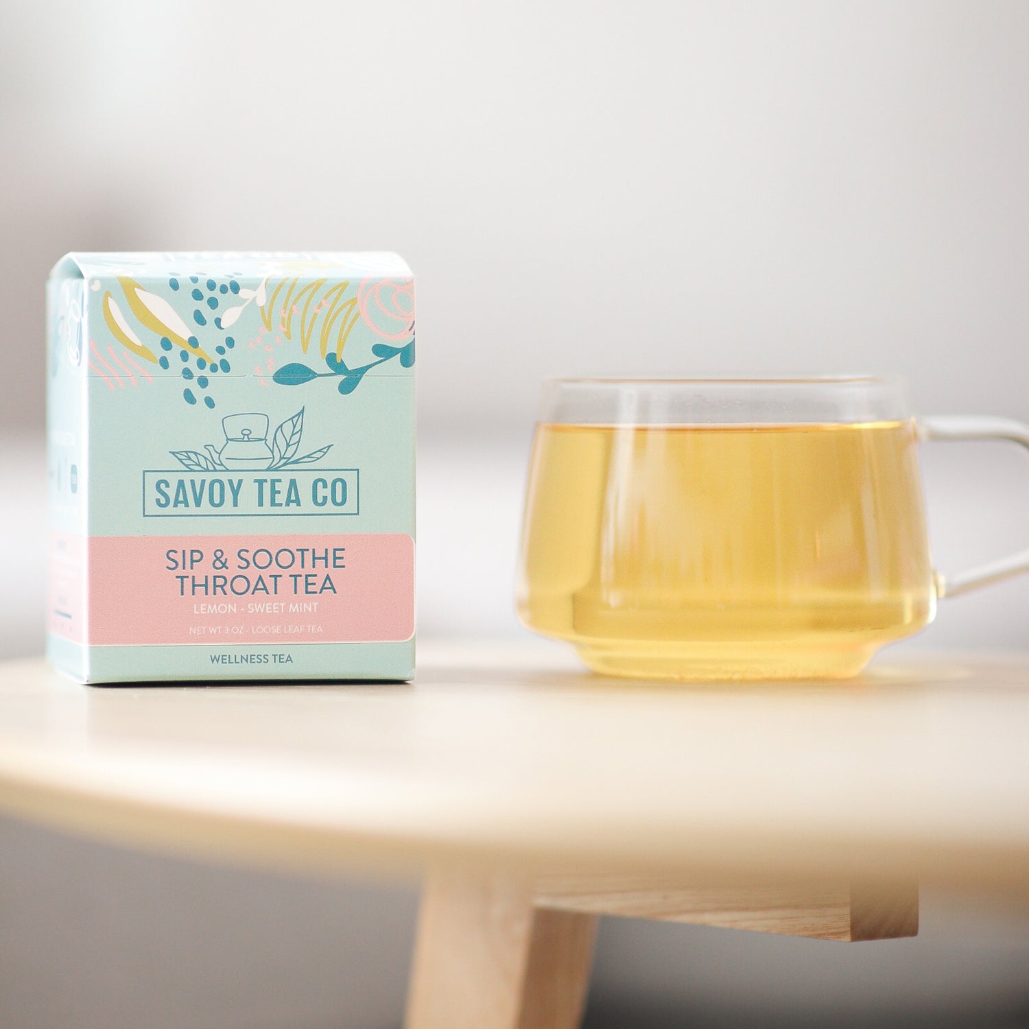 A box of Savoy Tea Co. Sip &amp; Soothe throat tea next to a clear mug filled with tea, placed on a wooden surface with a light background.